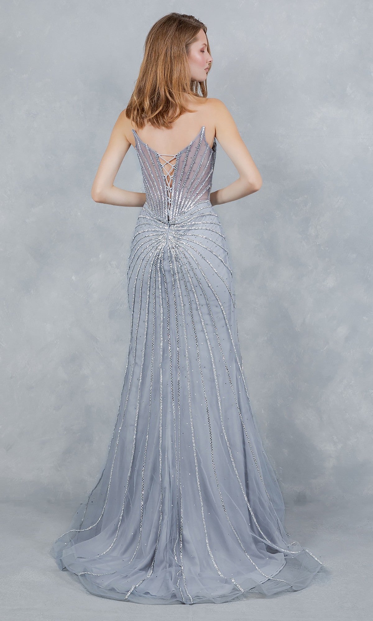 A woman faces away, wearing the Cinderella Couture 8576J—a floor-length silver-gray prom dress with a lace-up back and beaded linear patterns.
